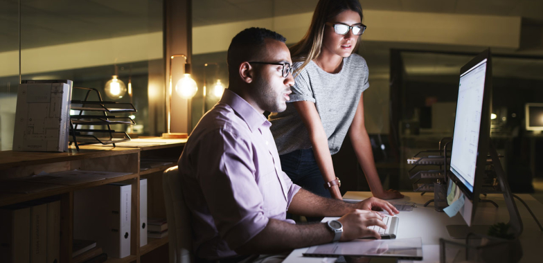 Male seated at desk and female standing, both looking at a computer screen