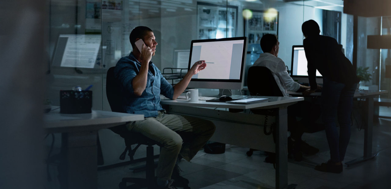 Man sitting at desk in an office, on the phone and pointing at his computer screen