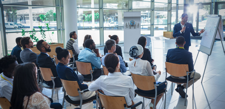Office workers sitting on chairs facing a man with microphone pointing at a white board