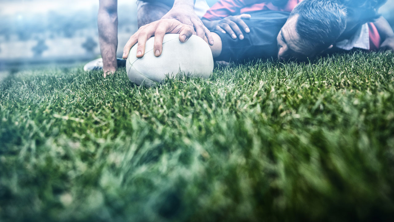 Rugby players in a scrum on the ground with one player's hand on the ball