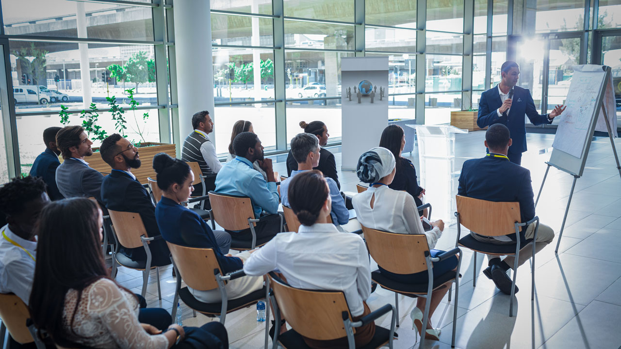 Office workers sitting on chairs facing a man with microphone pointing at a white board