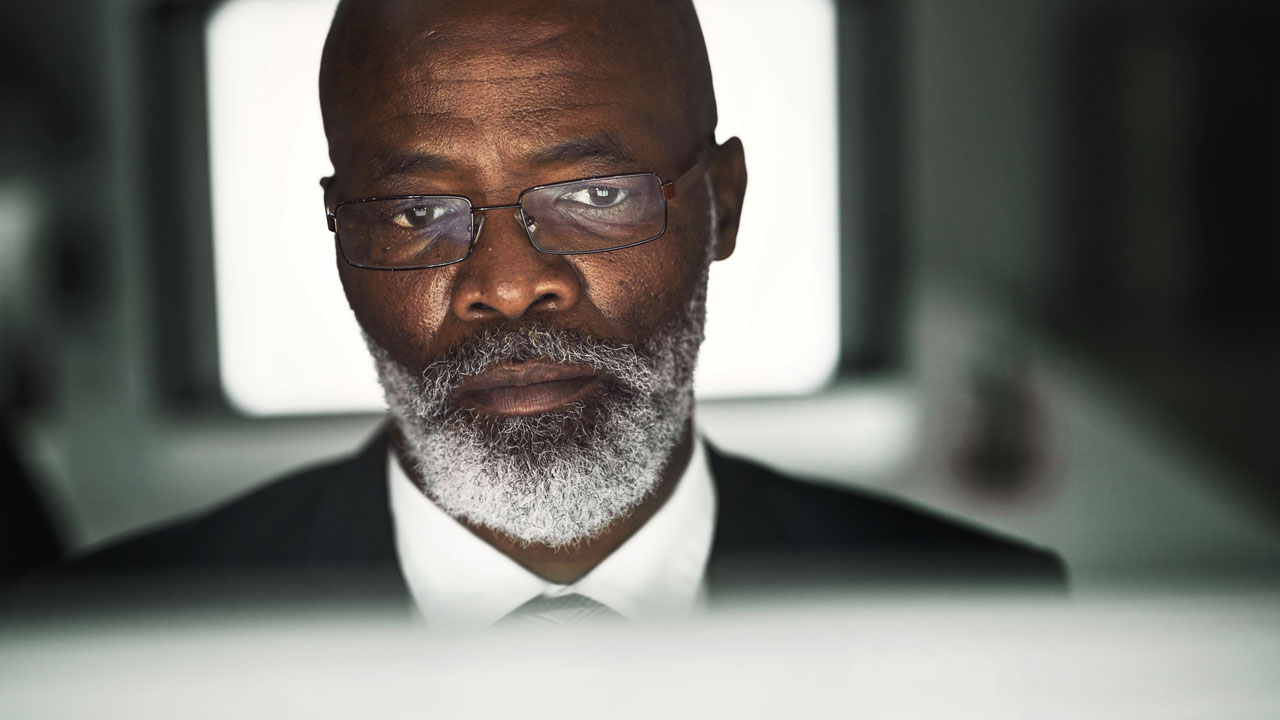 A man in glasses looking at a screen in the direction of the camera